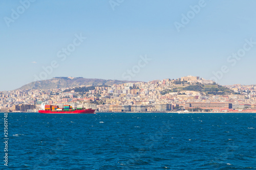 Red cargo ship with multi-colored containers in the Tyrrhenian Sea against the backdrop of the landscape of the city of Naples