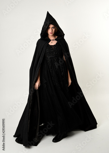 full length portrait of blonde girl wearing long black flowing cloak, standing pose with a white studio background.