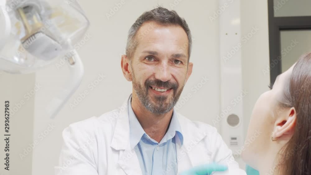 Charming male dentist smiling to the camera while working with patient. Attractive mature male dentist enjoying working at his clinic, checking teeth of a woman. Profession concept Charming male dentist smiling to the camera while working with patient. Attractive mature male dentist enjoying working at his clinic, checking teeth of a woman. Profession concept