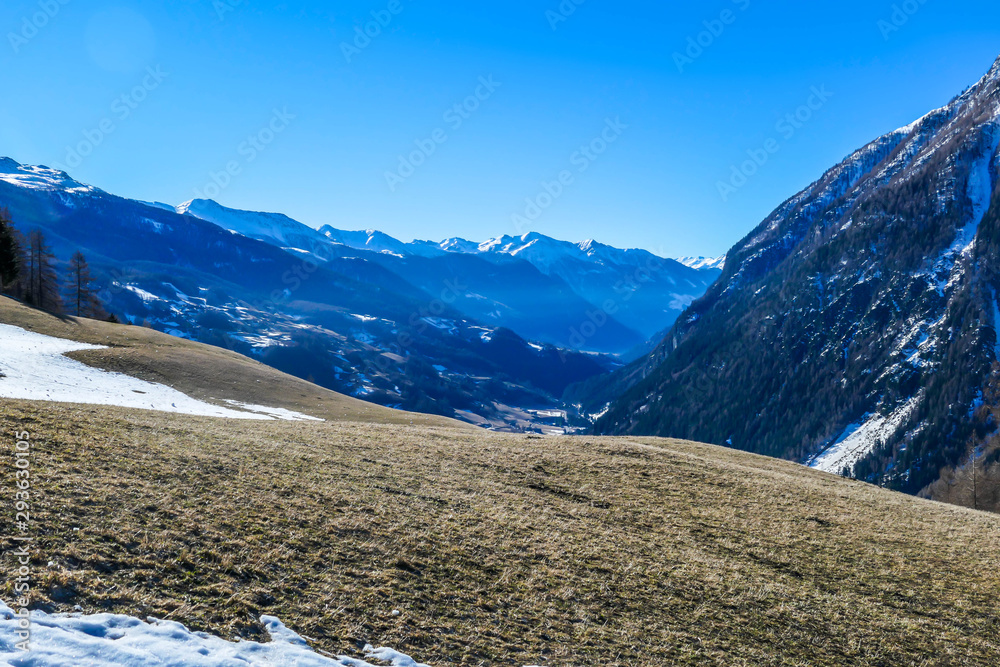 Spring slowly coming to tall Alps. Slopes are partially covered with snow. Grass is dried and colourless. Trees are golden. There is a little mist in the valley. Clear and sunny day.
