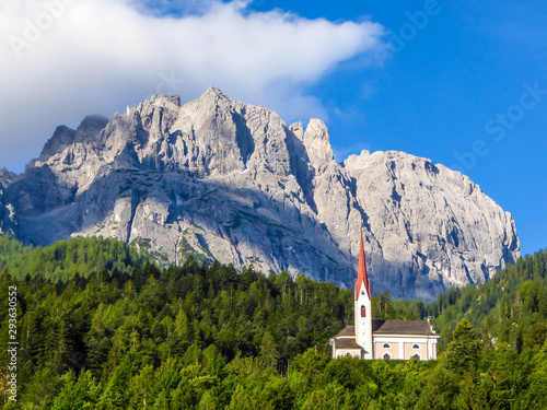 A church popping out of the forest, with a tall, sharp and rocky mountains behind. The church is build on a rock, making it taller than surrounding trees. Massive Alps in the back. Clear day. photo