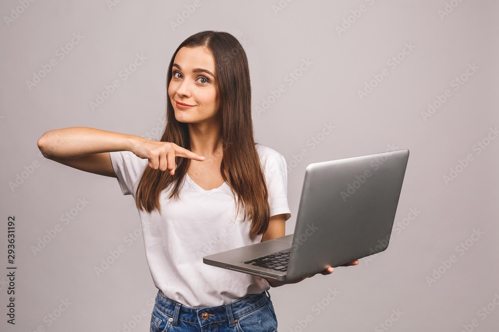 Naklejka premium Surprised happy brunette woman in casual showing blank laptop computer screen and pointing on it while looking at the camera isolated over grey background.