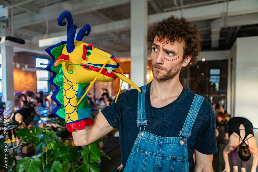 Obraz premium Diverse group of people in yoga class. A portrait of a funny Caucasian guy as he looks towards a hand puppet with a peculiar and curious face. Motivational man stands at front of yogic workshop.