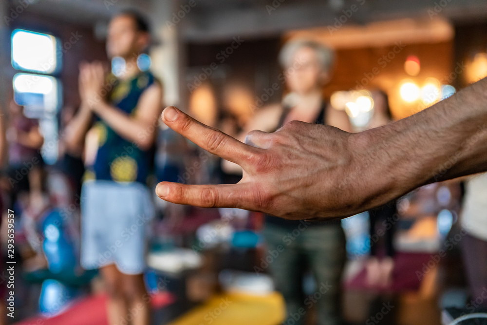 Diverse group of people in yoga class. A close-up view on the hand of a young man giving the v symbol at the front of a workshop dedicated to sun salutations. Blurred practitioners are seen behind.