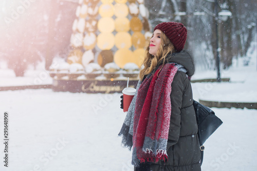 Beautiful smiling cute teenage girl with natural make up and light curly hair and red hat holds a cup with hot coffee or tea. Winter season. Christmas, New Year, winter holidays concept.