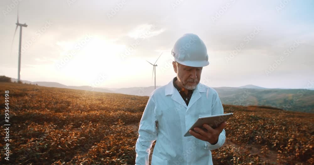 Mature caucasian engineer in hardhat and suit using tablet computer to check wind turbine system working at sunset on ecological station.