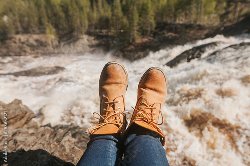 Woman's legs in blue jeans and light brown boots sitting on the edge near waterfall