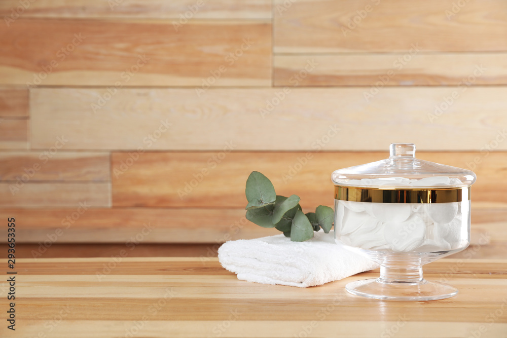 Decorative glass jar with cotton pads and towel on table against wooden background. Space for text Decorative glass jar with cotton pads and towel on table against wooden background. Space for text