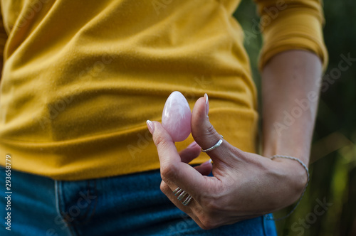 Adult girl in yellow shirt and blue jeans is holding pink quartz yoni egg for vumfit, imbuilding or meditation outdoors on her body background outdoors photo