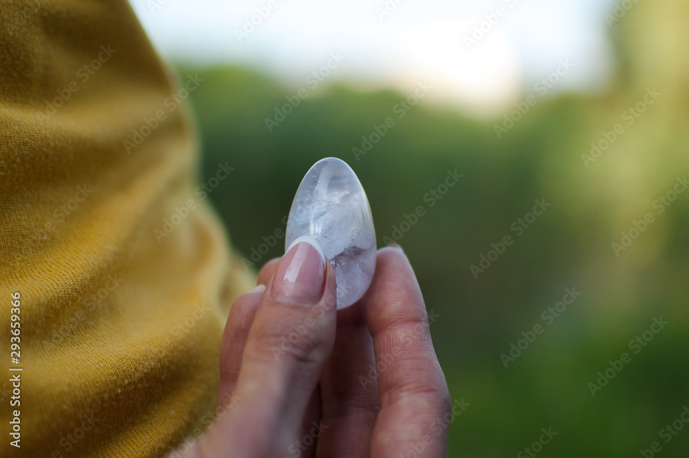 Adult girl in yellow shirt and blue jeans is holding transparent violet amethyst yoni egg for vumfit, imbuilding or meditation outdoors on her body background outdoors