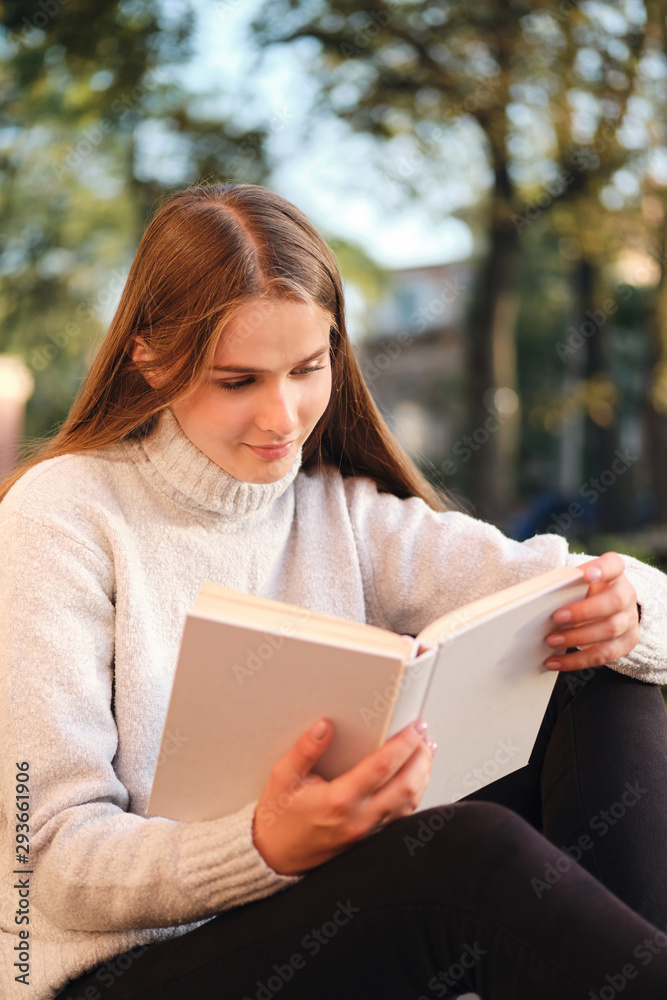 Obraz premium Young pretty student girl in cozy sweater dreamily reading book while studying outdoor