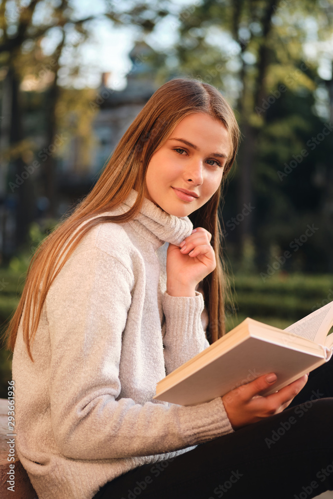 Obraz premium Young pretty smiling student girl in cozy sweater sensually looking in camera while studying with book outdoor