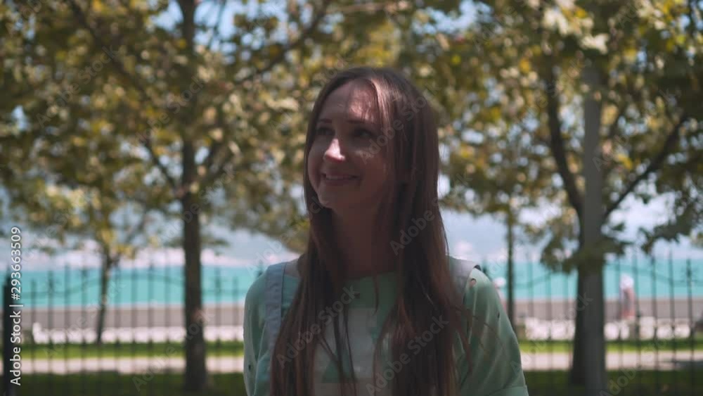A girl in white shorts and a T-shirt with long hair walks along the city promenade. A girl walks among green trees on the promenade against the background of the sea. A girl in white shorts and a T-shirt with long hair walks along the city promenade. A girl walks among green trees on the promenade against the background of the sea.