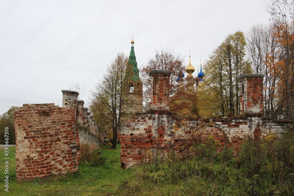 Fototapeta premium Path in old fence to cemetery, stone belfry and orthodox church with domes in cloudy autumn day