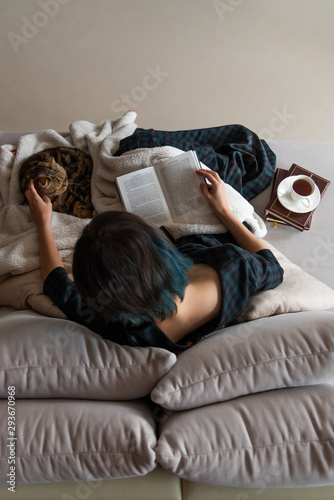 A reading woman sits on a sofa with a book and tea, caress a cat. top view.