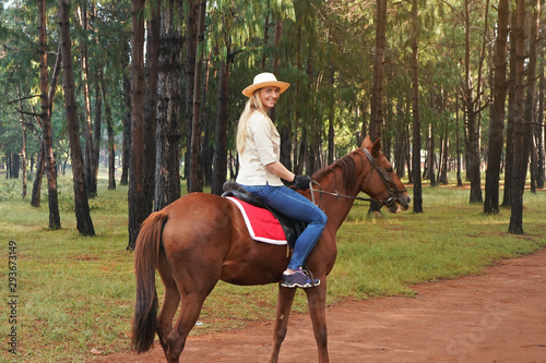 Young woman in shirt and straw hat, rides brown horse in the park, blurred trees in background