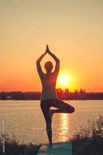 Vertical photo. A young girl is standing on the lake at sunset  doing yoga. Stands in a pose of tree a Sathi Yoga. Balance  harmony  balance  concentration  relaxation.