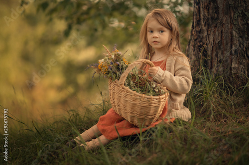 small girl sitting under the tree with wicker basket full of wildflowers and looking at the camera