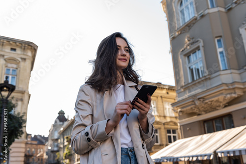 Smiling curly woman wearing trendy sunglasses walks down the central city street and uses her phone. Pretty summer woman in white jacket walks down the street looking at her mobile phone
