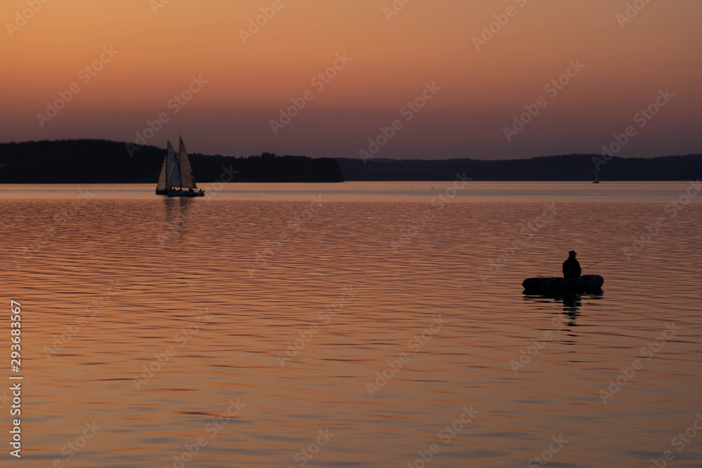 man fishing on a lake from the boat at sunset. silhouette of fishermen. yellow and orange sunset background. sailing boats in water at evening.