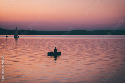 man fishing on a lake from the boat at sunset. silhouette of fishermen. yellow and orange sunset background. sailing boats in water at evening. © Natallia