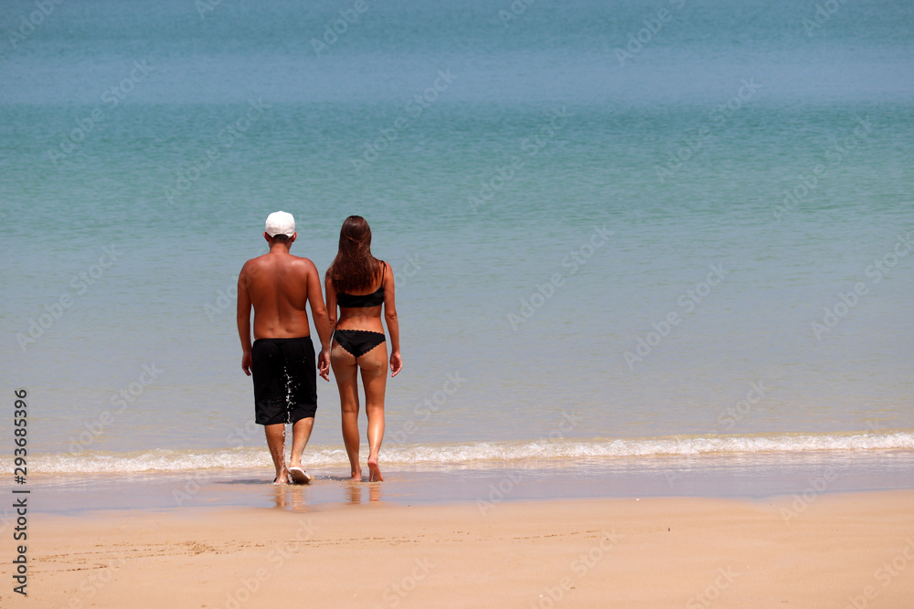 Couple going to swim in the tropical blue sea, colorful landscape. Sexy girl in black bikini and guy in shorts with sand on skin walk together, romantic leisure and beach activity