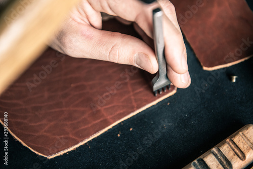 a young tanner on a desktop in a workshop punches holes for stitching on a piece of brown leather