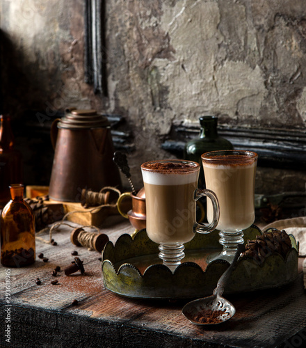 two tall glasses with layered coffee drink with foam and cocoa on top stands on vintage tray on rustic wooden table with copper pot, coffee beans opposite concrete wall, selective focus