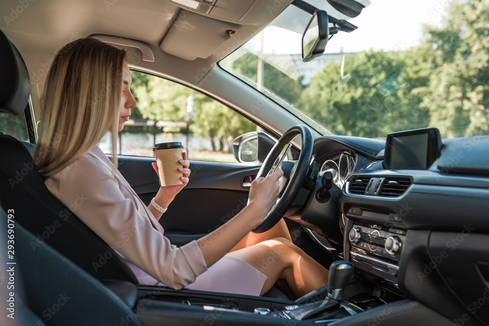 Fashionable stylish girl in car, in summer city woman holds mobile phone, cup with coffee tea, has a rest in parking lot, waiting in traffic jam on road. Snack and lunch while traveling.