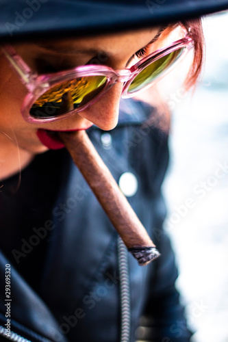 Closeup on the face of a beautiful young woman smoking a cigar during the sunset, she is looking down, has short hairstyle and a fashion style with sunglasses and a hat