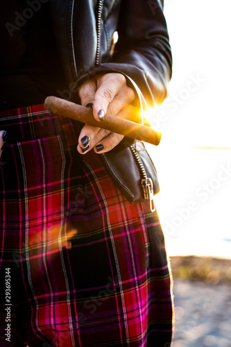 Extreme closeup on the hand of a young woman holding a cigar like a cigarette during the sunset on the beach, she is wearing a leather jacket