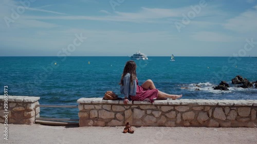 Beautiful woman sitting on the stone fence above town Antibes in South France, enjoying beautiful sea view. Distant plan 4k