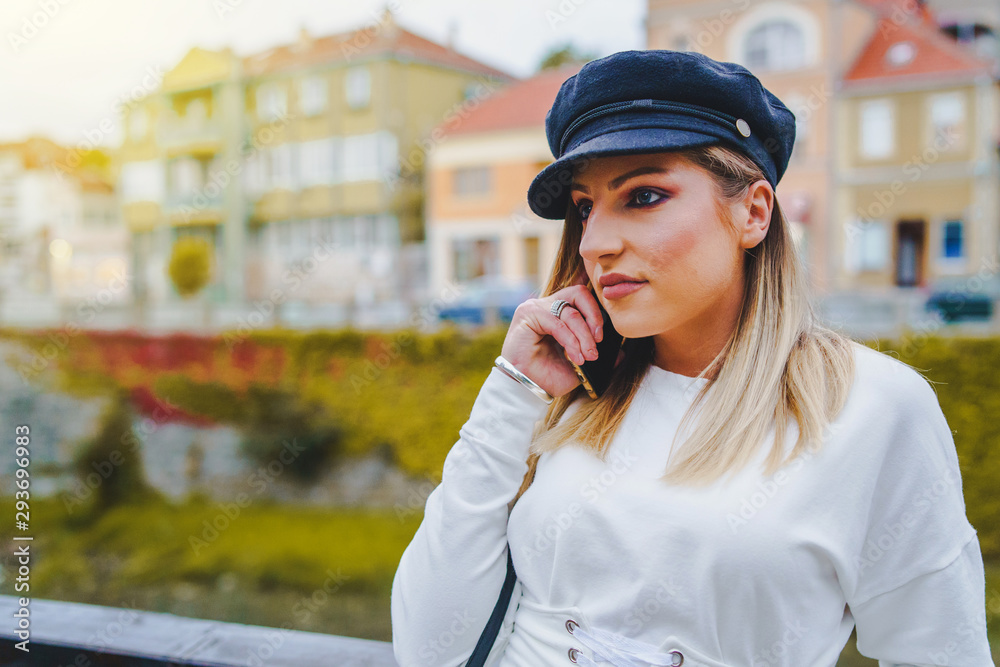 Young blond happy woman in white dress and black cap hat standing in town or city talking to the mobile phone using smart phone in autumn or spring day Young blond happy woman in white dress and black cap hat standing in town or city talking to the mobile phone using smart phone in autumn or spring day