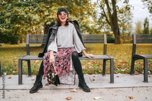Sincerely laughing female dressed fashion boho style colorful long dress, warm knitted sweater with black leather biker jacket and Beanie Hat with flap bag sitting on bench at autumn city park