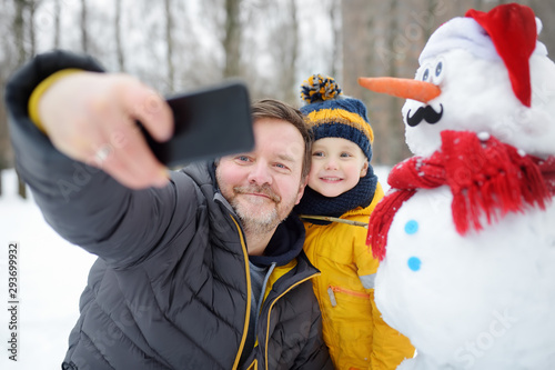 Little boy and his father taking selfie on background of snowman in snowy park. Active outdoors leisure with children in winter. photo