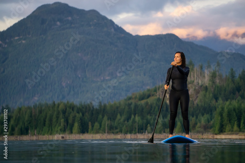 Adventurous Girl on a Paddle Board is paddling in a calm lake with mountains in the background during a colorful summer sunset. Taken in Stave Lake near Vancouver, BC, Canada.