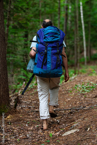 Diverse people enjoy spiritual gathering An older man is seen from behind, walking through a woodland trail with a large blue backpack during a camping retreat in nature.