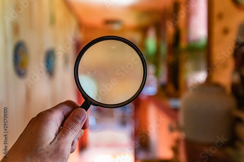 Indoor damp & air quality (IAQ) testing. A close up view on the hand of a man holding a magnifying glass indoors, a blurry background inside a hallway with wooden walls is seen.