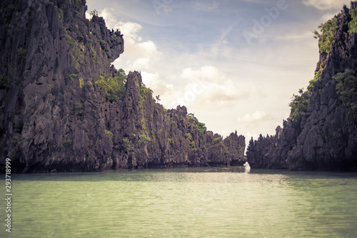 a long shallow lagoon is protected and surrounded by tall limestone rock walls with tall cliffs to protect the small beach from wind and water erosion with a distant cave exit to the sea