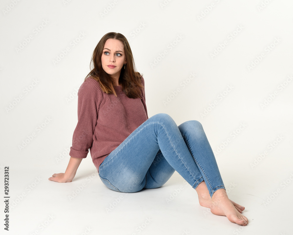 full length portrait of brunette woman wearing jeans and pink jumper. seated pose with a white studio background.