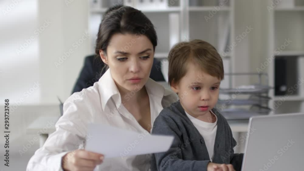 Businesswoman working with documents and reading notes in notepad at office desk while little son sitting on her knees and playing on laptop