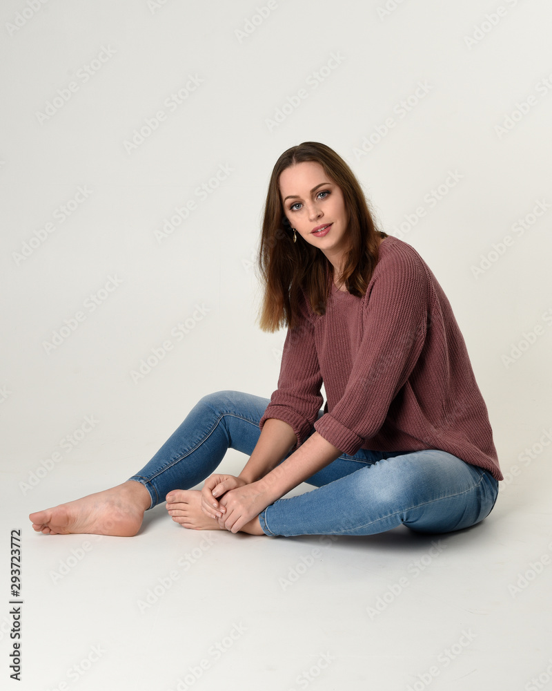 full length portrait of brunette woman wearing jeans and pink jumper. seated pose with a white studio background.