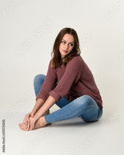 full length portrait of brunette woman wearing jeans and pink jumper. seated pose with a white studio background. © faestock