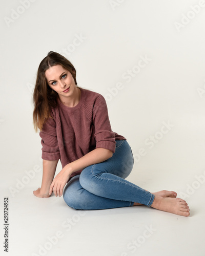 full length portrait of brunette woman wearing jeans and pink jumper. seated pose with a white studio background. © faestock
