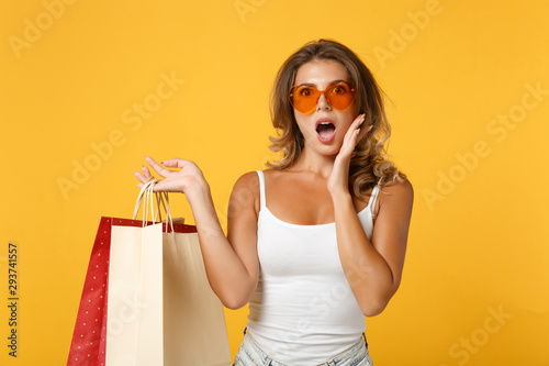 Shocked young woman girl in eyeglasses, light casual clothes posing isolated on yellow orange background. People lifestyle concept. Mock up copy space. Hold package bag with purchases after shopping.