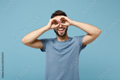 Young cheerful smiling man in casual clothes posing isolated on blue wall background in studio. People lifestyle concept. Mock up copy space. Holding hands near eyes, imitating glasses or binoculars.