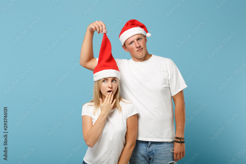 Obraz premium Confused young Santa couple friends guy girl in Christmas hat posing isolated on pastel blue background. Happy New Year 2020 celebration holiday concept. Mock up copy space. Covering mouth with hand.