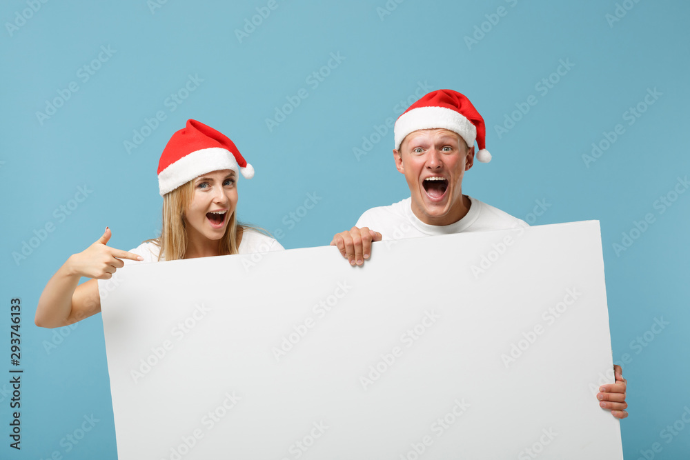 Joyful young Santa couple friends guy girl in Christmas hat isolated on blue background. Happy New Year 2020 celebration holiday concept. Mock up copy space. Holding big white empty blank billboard. Joyful young Santa couple friends guy girl in Christmas hat isolated on blue background. Happy New Year 2020 celebration holiday concept. Mock up copy space. Holding big white empty blank billboard.