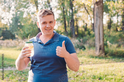 Man in casual wear posing over green city park on sunny warm day, holding cup of lemonade, giving thumbs up and looking to camera with smile. Vitamins and regular life concept