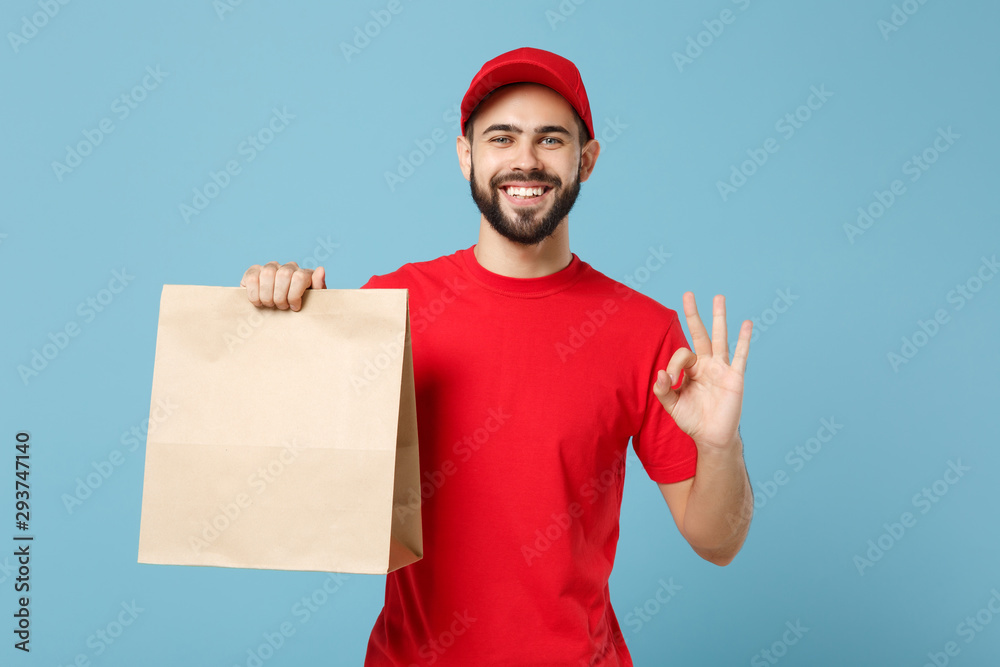Delivery man in red uniform hold craft paper packet with food isolated on blue background, studio portrait. Male employee in cap t-shirt print working as courier. Service concept. Mock up copy space. Delivery man in red uniform hold craft paper packet with food isolated on blue background, studio portrait. Male employee in cap t-shirt print working as courier. Service concept. Mock up copy space.